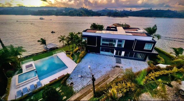 Golden-hour aerial view of a black-and-white lakefront villa with terraced pools, palm landscaping and the reservoir beyond.