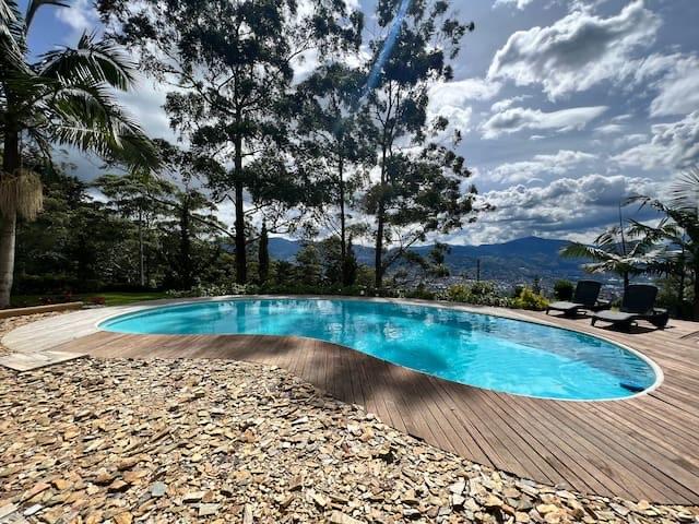 Kidney-shaped pool on a wooden deck with loungers, tall pines and panoramic views of the Medellín valley and mountains.