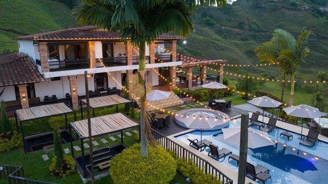 Elevated dusk view of a white two-storey villa with clay tile roof, string lights, pergolas, a round jacuzzi and a large pool with umbrellas.