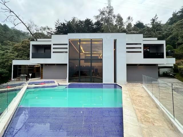 Daytime view across the blue-tiled pool to the white and gray modern house with a double-height glass facade.