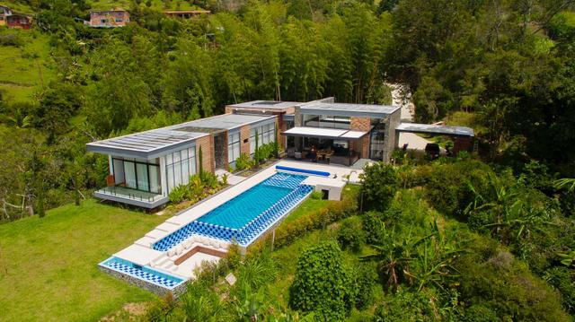 Aerial daytime view of a low flat-roofed modern villa with a long checkerboard-edged pool, large lawn and surrounding forest.
