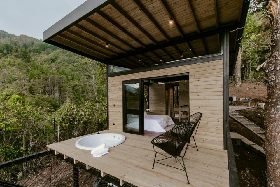 Timber cabin with a covered wooden deck, an oval outdoor tub and black wire chairs facing forested mountains.