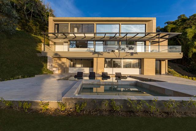 Daytime exterior of Casa Cielo with a beige stone facade, glass-railed terrace and pool surrounded by trees.