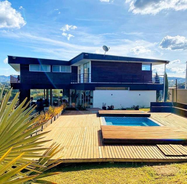 Daytime view of a two-storey house with dark wood-clad upper floor and white glazed lower floor, behind a wooden deck with a small rectangular pool.