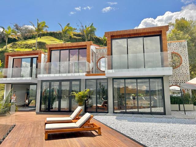 Daytime front view of a white two-storey villa with wood-framed upper volumes, glass balconies and a wooden deck with loungers.