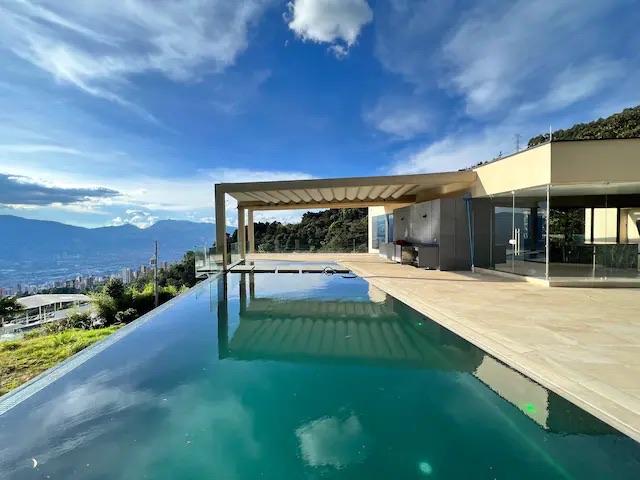 Daytime infinity pool with a covered pergola terrace and glass-walled modern house overlooking the Medellín valley.