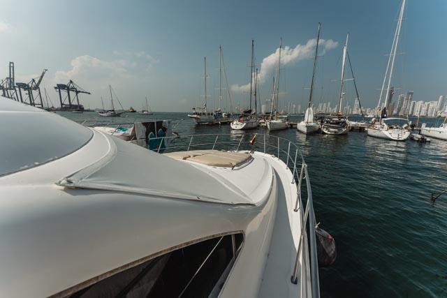 White hull and side deck of the yacht at anchor among sailboats, with port cranes and the Cartagena skyline in the distance