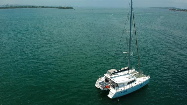 Aerial stern-quarter view of a white sailing catamaran with twin hulls, mast and a tender on the aft davits