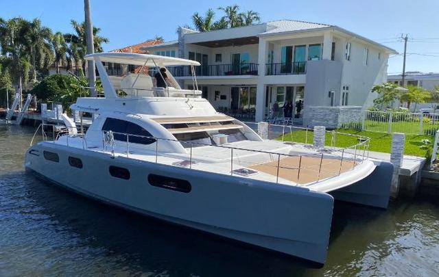 Bow-quarter view of a white and grey twin-hulled powercat moored beside a waterfront house, with a covered flybridge and brown foredeck panels.