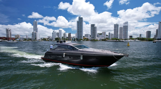 Dark grey sport yacht with a red waterline cruising at speed past the Cartagena skyline under a bright cloudy sky