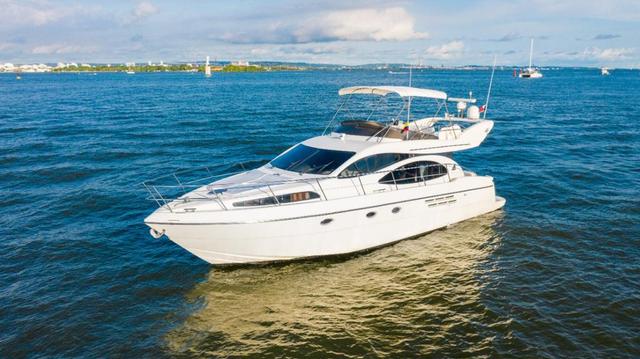 Haya, a white flybridge motor yacht with a canopy-covered upper deck, at anchor on blue water with the Cartagena shoreline behind