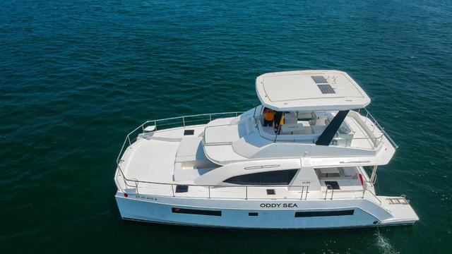 Aerial side view of Oddy Sea, a white powercat with visible ODDY SEA hull lettering, foredeck cushions and a covered flybridge.