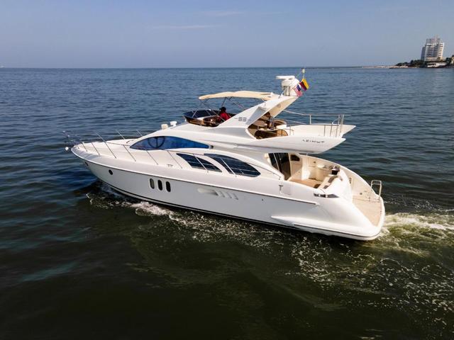 White Azimut flybridge yacht cruising with guests on the upper deck, a Colombian flag at the stern and the Cartagena shoreline behind