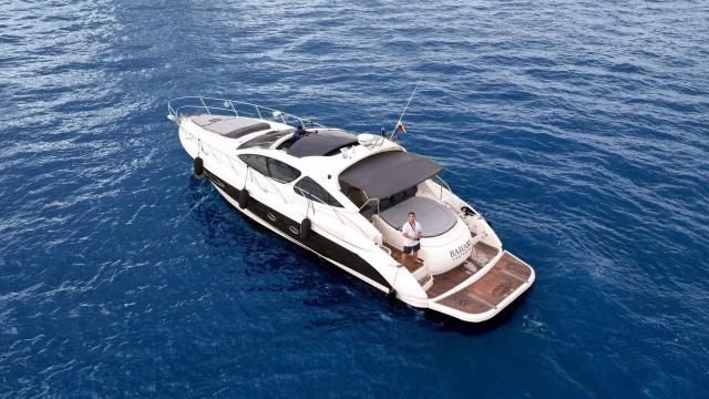 Aerial view of Bahari at sea, its name on the stern, with a teak swim platform, black canopy and crew on deck